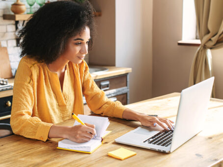 Woman in a yellow shirt sitting on the kitchen table, writing down things from the laptop in front of her