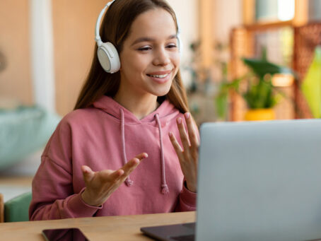 Girl in pink sweatshirt with headphones speaking online on laptop in front of her