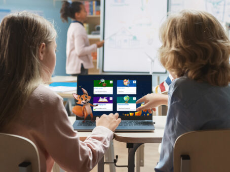 Two kids from behind pointing to a laptop, with a teacher on board in the background